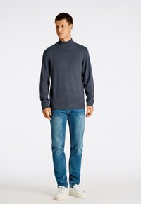 Mörkgrå polotröja med långa ärmar, kombinerad med ljusblå jeans och vita sneakers, presenterad mot en enfärgad bakgrund.