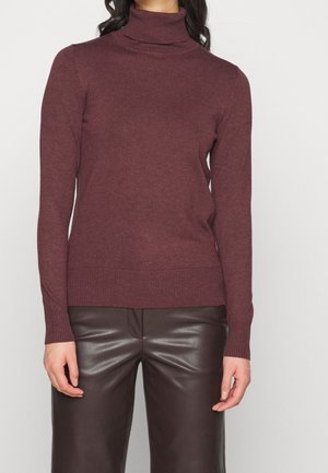 Kobieta w dopasowanym bordowym swetrze z wysokim kołnierzem i ciemnobrązowych skórzanych spodniach, stojąca na tle jednolitej białej ściany.