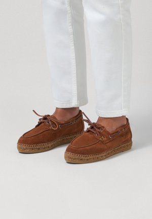 Brune mokkasinstil-sko i ruskind med reb såler, båret med hvide korte bukser på en lys baggrund.