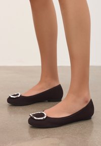 Des ballerines ensuédées marron foncé avec un bout arrondi et un accent métallique argenté à l'avant. Talon bas et texture lisse visible.