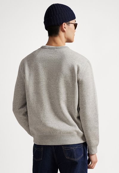 Siva bombažna jopa z rebrastim okroglim ovratnikom in manšetami, nošena s temno modrimi kavbojkami. Oseba nosi mornarsko modro pleteno kapo in sončna očala.