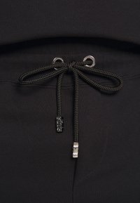 Gros plan sur un cordon noir noué en forme de nœud sur un tissu noir, avec des embouts métalliques et des œillets gravés de texte.