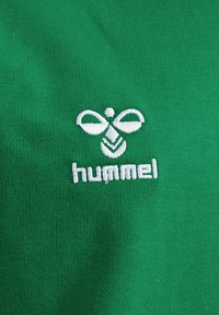 Tessuto di cotone verde con una superficie testurizzata presenta un logo ricamato bianco di due ali con la parola "hummel" sottostante.