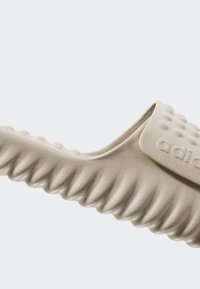 Beige liukusandalai su tekstūruota, bangų rašto dizainu, turintys platų dirželį ir mažus iškilusius taškus sukibimui. Pagaminti iš lankstaus medžiagos.