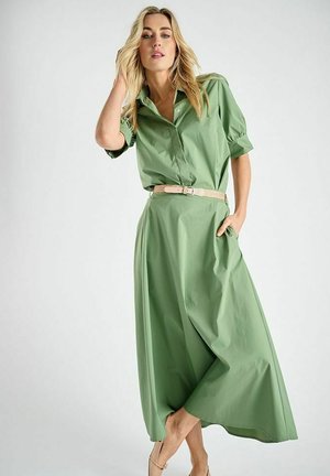 Vrouw draagt een lichtgroene jurk met knopen en een beige riem, poseert met de ene hand in haar zak en de andere hand haar haar aanrakend.