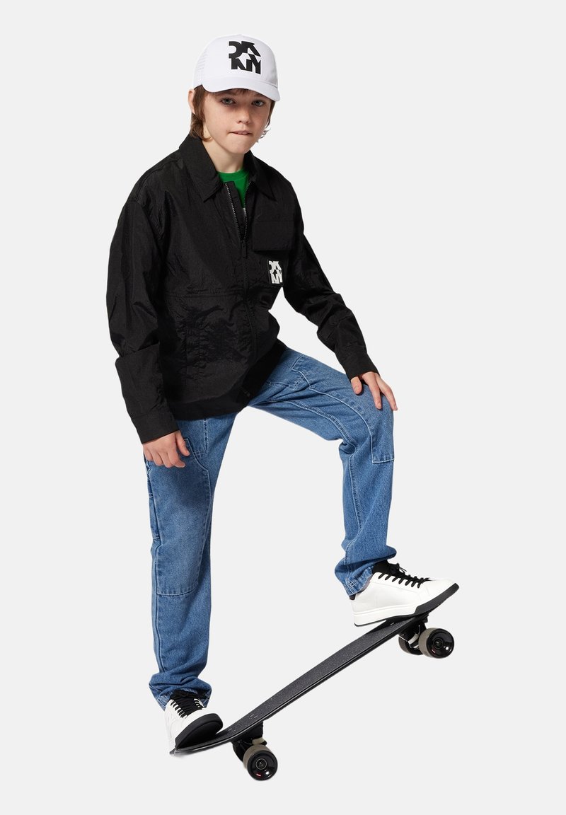 Veste zippée noire avec poche avant, pantalon en denim bleu, baskets blanches et casquette blanche avec un logo noir, posée sur un skateboard.