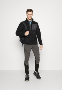 Black fleece pullover with a gray chest panel, paired with gray and black tapered pants featuring reflective accents; accessorized with a black backpack.