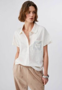 Polo à manches courtes blanc avec une texture subtile, doté d'une poche avant ornée d'un motif en maille. Assorti à un pantalon beige à motif.
