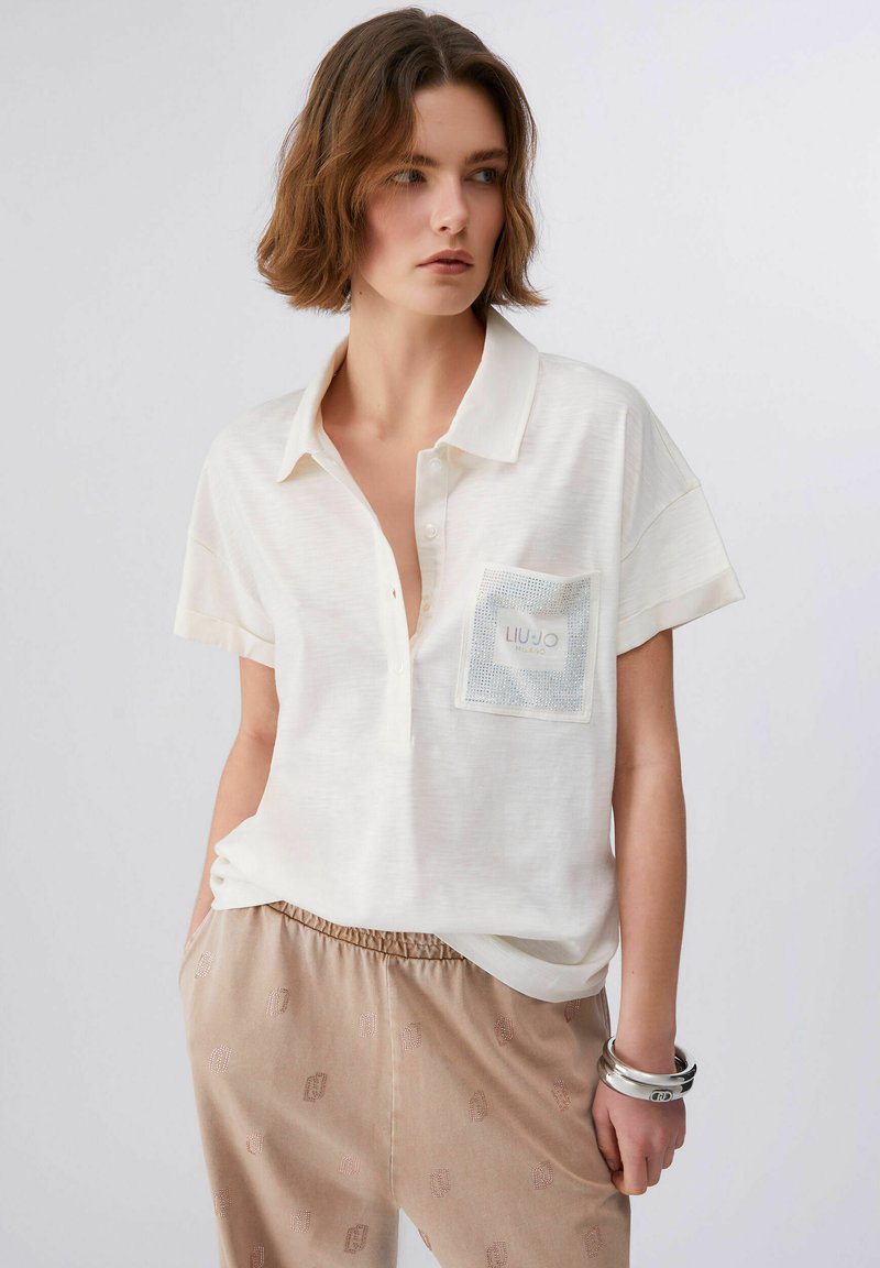 Polo à manches courtes blanc avec une texture subtile, doté d'une poche avant ornée d'un motif en maille. Assorti à un pantalon beige à motif.