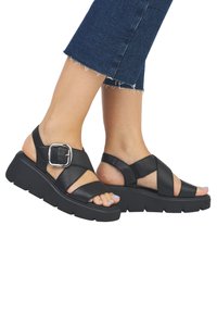 Sandales en cuir noir avec une semelle épaisse, dotées de lanières croisées, d'une grande boucle argentée et d'un design ouvert au niveau des orteils.