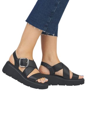 Black leather sandals with a chunky platform, featuring cross straps, a large silver buckle, and an open toe design.