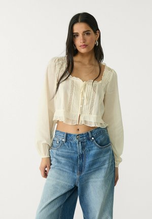 Cream-colored, semi-sheer blouse with long sleeves, ruffled hem, and textured front panel. Paired with light blue high-waisted jeans.