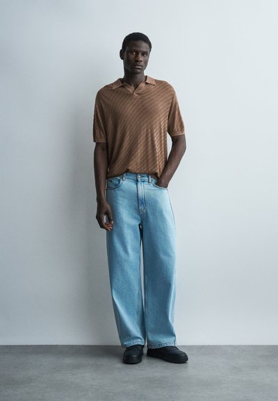 Un homme se tient contre un mur gris clair uni, portant une chemise marron ample à manches courtes texturée, un jean large bleu clair et des chaussures noires.