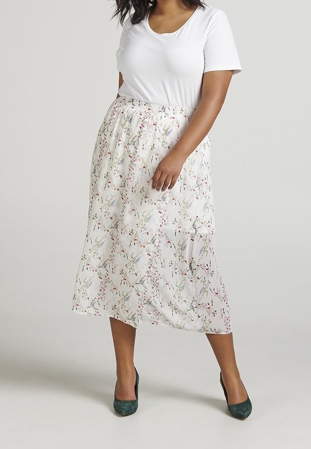Femme portant un haut blanc à manches courtes, une jupe midi blanche à petit imprimé floral, et des chaussures à talons hauts vert foncé, debout devant un fond uni.
