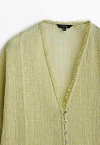 Lichtgroene linnen blouse met een V-hals, rafelige randen en vier ronde knopen aan de voorkant. Getextureerde stof met een zachte glans.
