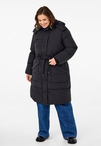 Zwarte pufferjas met een ceintuur in de taille, een capuchon en knoopsluiting. Voorzien van grote zakken en een gewatteerde textuur. Gecombineerd met wijd uitlopende blauwe spijkerbroeken.