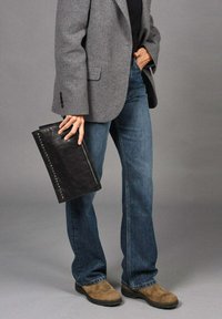 Clutch de cuero negro con acentos de tachuelas, sostenido en la mano. Blazer gris sobre una blusa negra, combinado con pantalones vaqueros de campana azules y botas marrones.