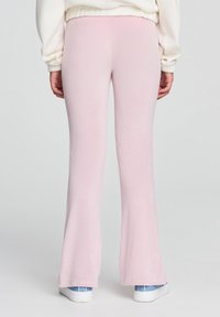 Osoba ubrana w jasnoróżowe, żebrowe spodnie w fasonie dzwonów oraz białe trampki, widoczna od tyłu na jednolitym tle.