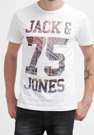 T-shirt blanc avec "JACK & JONES" et un grand numéro 75 affichant une impression de rue de la ville et de véhicules, porté par une personne avec un tatouage de boussole sur l'avant-bras gauche.
