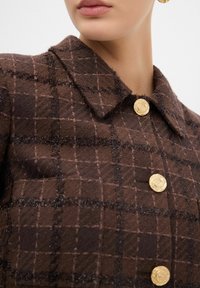 Giacca marrone con texture e motivo a quadri, colletto e due bottoni decorativi dorati. Il tessuto ha un aspetto morbido e peloso.