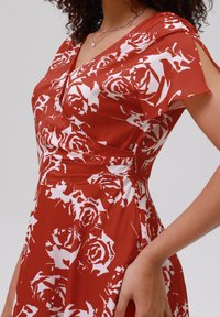 Robe rouge à motifs de roses blanches, avec un décolleté en V et des manches courtes. Confectionnée en tissu lisse, elle présente un buste ajusté et une jupe évasée.