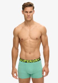 Boxer shorts de algodón verde con una banda negra que presenta texto amarillo. Los shorts tienen un diseño ajustado y una textura suave.