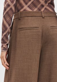 Καφέ, υφασμάτινο παντελόνι με προσαρμοσμένη εφαρμογή, που διαθέτει δύο πίσω τσέπες και λεία μέση. Το ύφασμα έχει ένα διακριτικό σχέδιο.