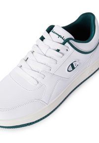 Weiße Sneakers mit niedrigen Schaft und dunkelgrünen Akzenten an der Umrandung, den Schnürsenkeln und der Sohle, ausgestattet mit dem Champion-Logo an der Seite und auf der Zunge.