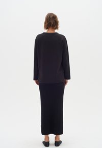 Top lungo nero con maniche lunghe e scollatura rotonda, abbinato a una gonna maxi nera aderente. Il tessuto sembra liscio e leggero. Calzature nere.