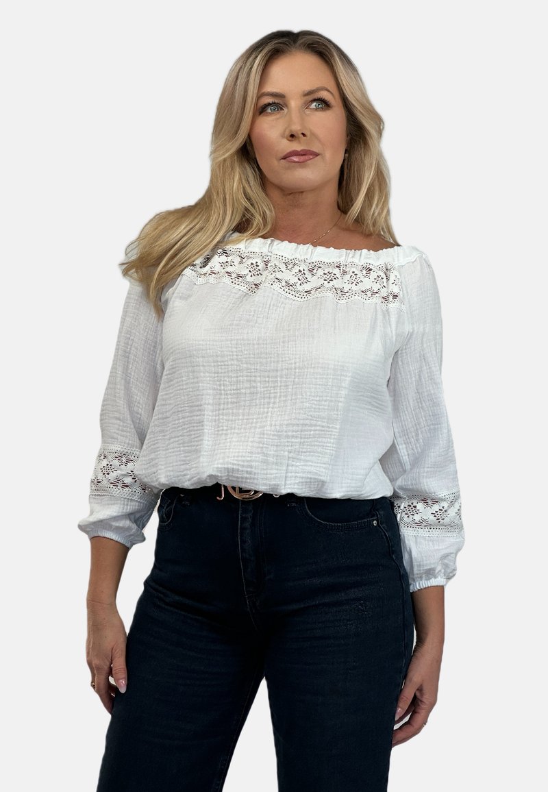 Biała bluzka z długim rękawem, wykonana z teksturowanej tkaniny, z koronkowymi detalami na ramionach i rękawach, w połączeniu z ciemnoniebieskimi dżinsami.