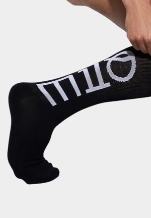 Ręka ściągająca czarną skarpetę z dużym białym pionowym napisem na łydce na jasnym, jednolitym tle.