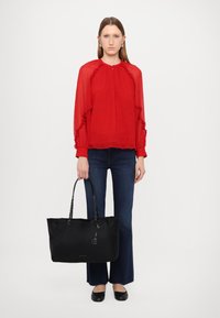 Borsa a tracolla nera con manici a catena, etichetta logata, tessuto morbido. Indossata con una blusa rossa brillante a ruffle e jeans a zampa d'elefante blu, scarpe piatte.