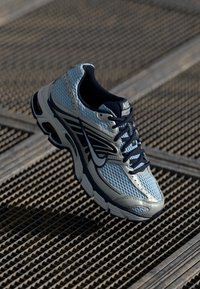 Svetlomodré a námornícky modré bežecké tenisky Nike s priedušným zvrškom, ktoré sa vznášajú nad kovovou mriežkovou podlahou.