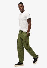 Olivengroene cargo pants met meerdere zakken, een trekkoord in de taille en lichte katoen, gecombineerd met een eenvoudig wit T-shirt en zwarte sneakers.