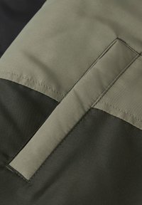 Gros plan sur un tissu vert foncé avec une bande cousue vert clair et un détail de patch rectangulaire sur un vêtement ou un équipement.