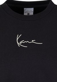 Svart hettegenser med rund hals, har en hvit brodert logo i kursiv stil. Mykt stoff med en jevn tekstur.