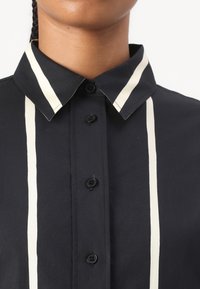 Black shirt with a pointed collar and white vertical stripes. Features black buttons along the front and smooth fabric texture.