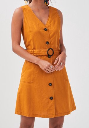 Kvinde iført ærmeløs sennepsgul knap-i-front-kjole med bælte og sort rundt spænde, stående med hænderne foldet.
