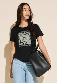 Černé tričko s grafickým potiskem, na němž je nápis "SLOW VIBES". Model drží černou koženou tašku. Světle modré džíny, volný střih.