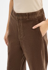 Brązowe spodnie w kant z szerokimi nogawkami, o teksturowanej powierzchni, z przednimi kieszeniami i małym metalowym guzikiem w talii.