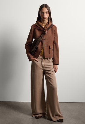 Mujer con chaqueta marrón, suéter mostaza, pantalones anchos beige, zapatos con estampado de leopardo y un bolso marrón cruzado con una bufanda alrededor del cuello.