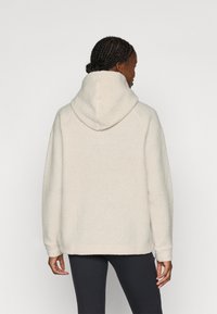 Fleece kapuce off-white krāsā ar lielu kapuci, kritiem pleciem un ribotām piedurknēm, ar teksturētu virsmu un brīvu piegulumu.