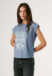 Zila kokvilnas īsā kreklā ar īsām sarullētām piedurknēm, ar baltu ziedu grafiku un zīmola logotipu priekšā. Apvienots ar tumšiem džinsiem.