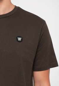 Dunkelbraunes kurzärmliges T-Shirt mit einem kleinen schwarzen runden Aufnäher, der ein weißes "W" auf der linken Brustseite zeigt.