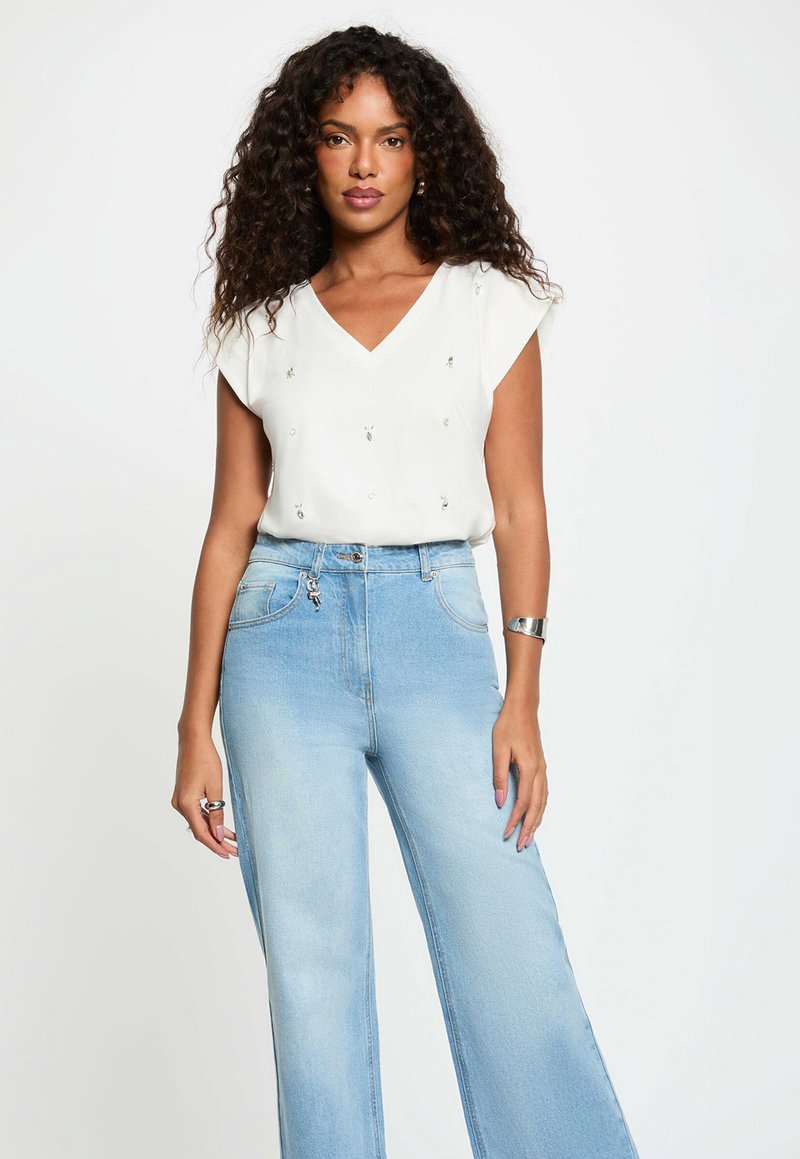 Woman with curly hair wearing a white short-sleeve blouse with small embellishments and high-waisted light blue wide-leg jeans.