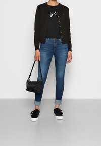 Cardigan noir porté sur un t-shirt noir avec texte blanc, jean skinny bleu avec revers roulés, sac noir et baskets noires avec fermetures velcro.