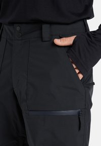 Zwarte waterdichte broek met een platte tailleband met twee knopen, een ritszijzak en een gladde textuur. Rekbare stof voor bewegingsvrijheid.