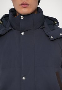 Giubbotto imbottito blu navy con colletto alto, bottoni a pressione e chiusura con zip, dalla texture liscia e dettagli in contrasto marrone.