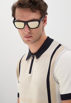 Giovane uomo che indossa occhiali da sole rettangolari neri e una polo a maniche corte beige e nera in maglia con colletto con cerniera.