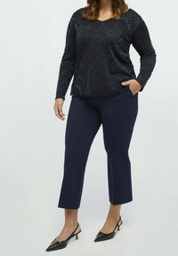 Långärmad svart topp med teal paisleymönster, parat med marininspirerade cropped byxor. Modellen bär svarta klackskor och har händerna i fickorna.